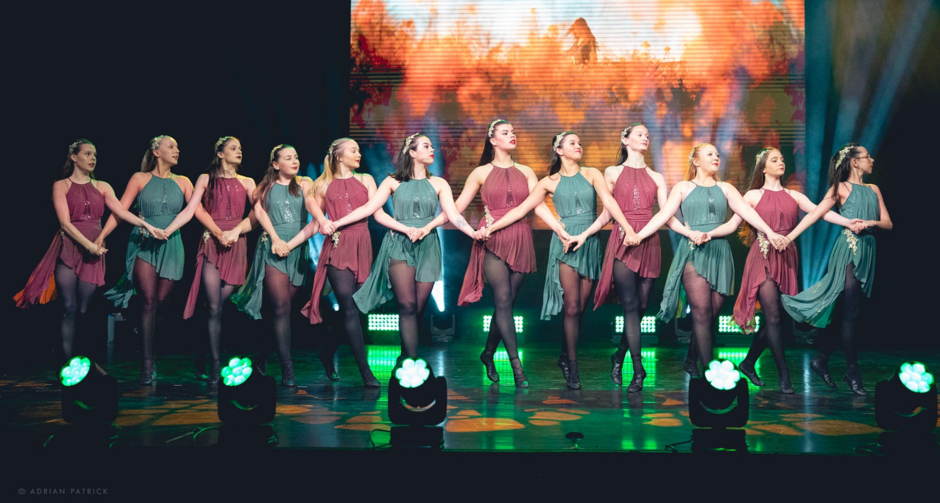Irish dancers in green and burgundy costumes holding hands across the stage